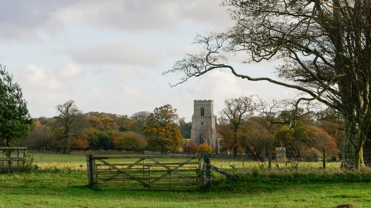 A rural autumn landscape with a wooden farm gate opening onto a grassy field. In the distance, a stone church tower rises among trees displaying autumn colours. Large, trees frame the scene, while a cloudy sky casts soft light across the countryside.